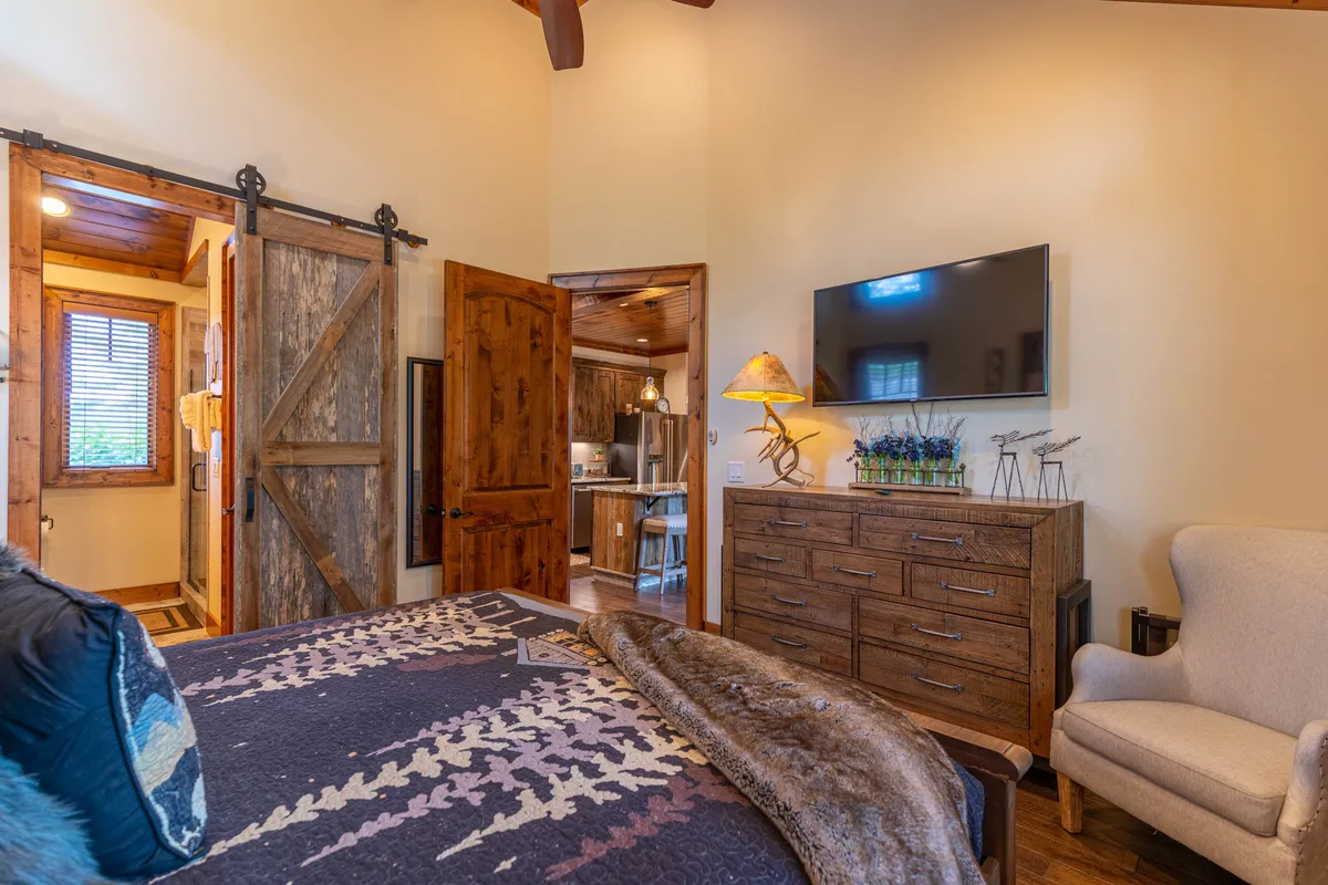 A warmly decorated king suite with a bed dressed in a patterned blanket, a wooden dresser, wall-mounted TV, armchair, and a sliding barn door leading to the ensuite. The combination of comfort and style highlights the suite's inviting ambiance.
