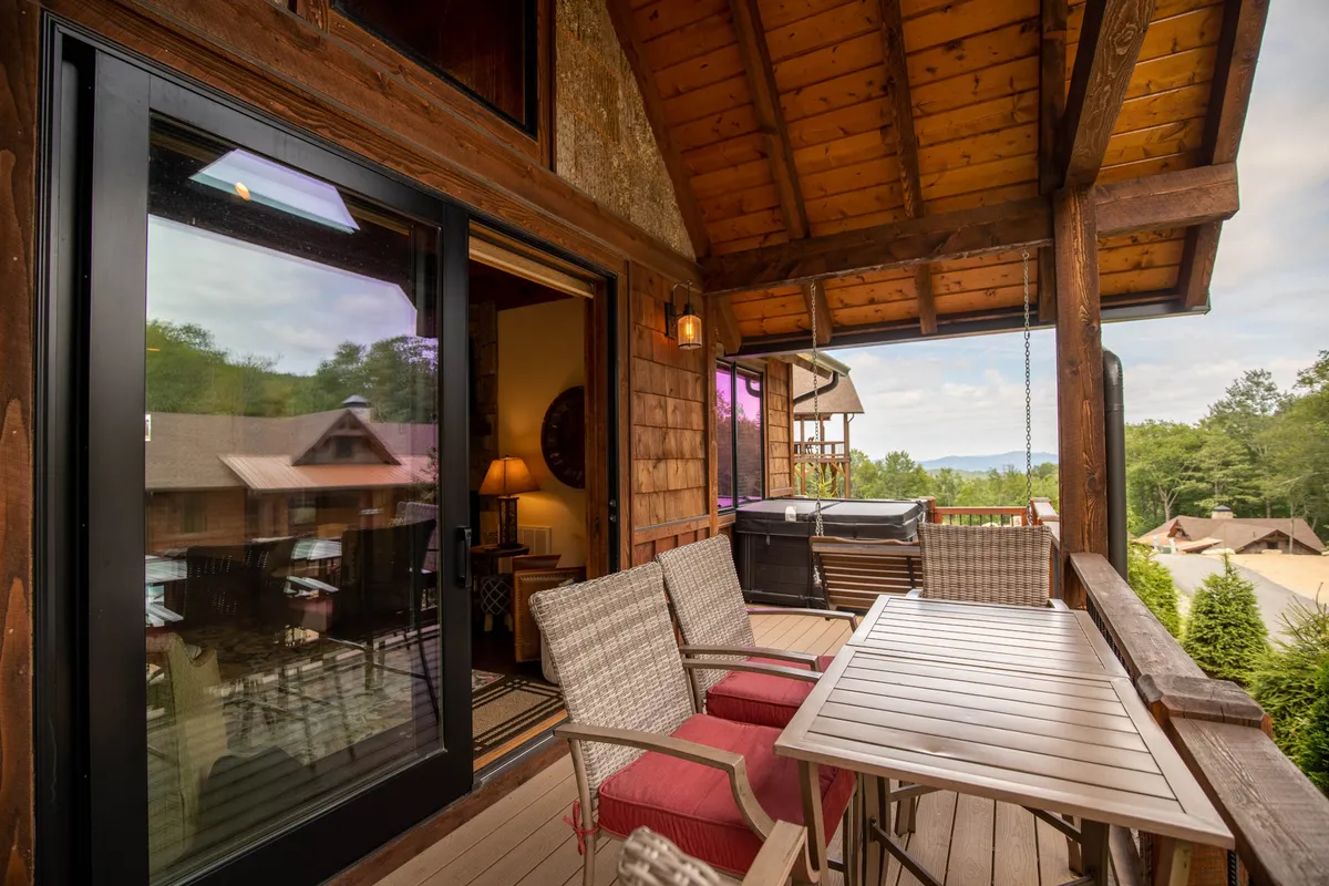 This exterior image showcases a cozy balcony furnished for socializing, featuring a private hot tub overlooking beautiful mountain views. The scene is enhanced by warm interior lighting spilling out onto the deck, inviting guests to enjoy both relaxation and companionship under the open sky.