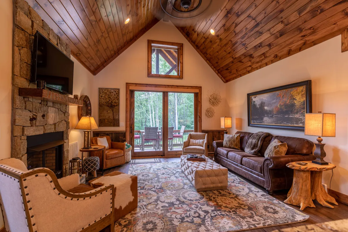 A bright, welcoming living room with a plush sofa, large windows drawing in mountain views and sunlight, a central rug, and a glowing gas fireplace that serves as the room's focal point. The setting is ideal for relaxing, socializing, or enjoying a peaceful moment by the fire.