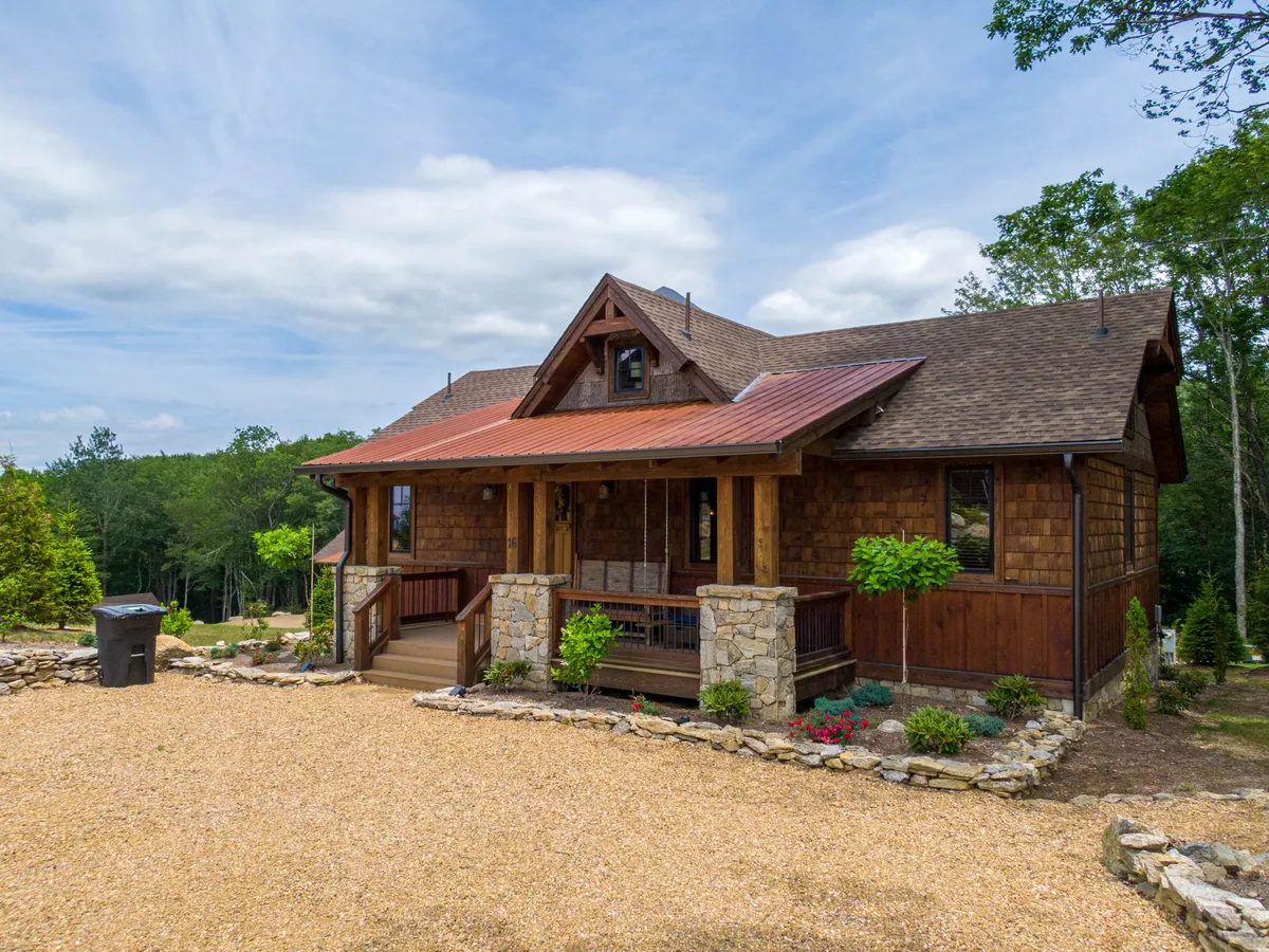 The image showcases the inviting wooden porch of a cozy cabin set on a sturdy rock foundation, surrounded by a beautifully landscaped yard and topped with a sleek metal roof. The porch overlooks serene mountain scenery, perfect for enjoying sunset views or a peaceful morning coffee immersed in nature.