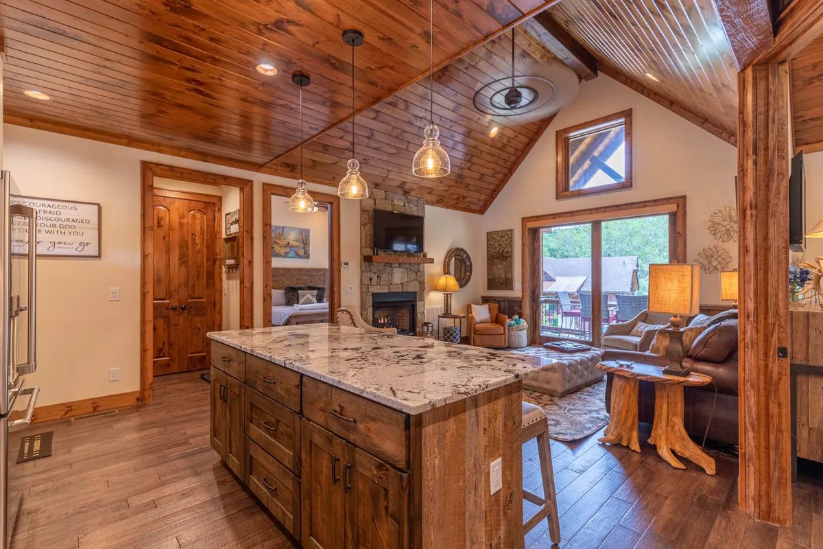 An open-concept living and kitchen area featuring a spacious island countertop, a cozy fireplace, wood-paneled ceilings, and expansive windows letting in abundant natural light. There's plenty of seating for gathering, and the scene captures both the kitchen and living areas in one cohesive space.