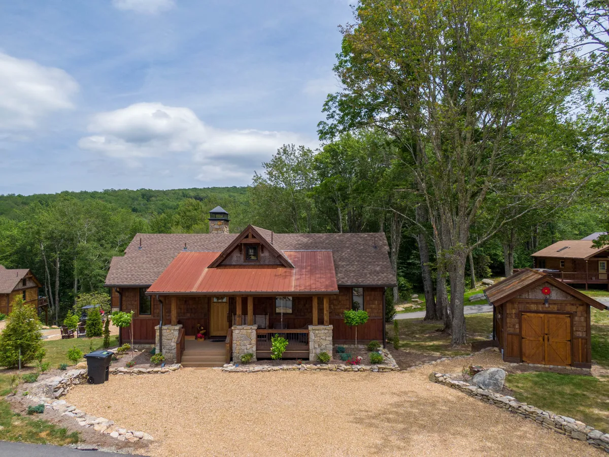 A warm, inviting view of a wooden house with a red roof, surrounded by grand trees and a stone pathway leading to the porch. The setting hints at both privacy and proximity to the natural amenities, with areas for family gatherings and easy access to the outdoors.