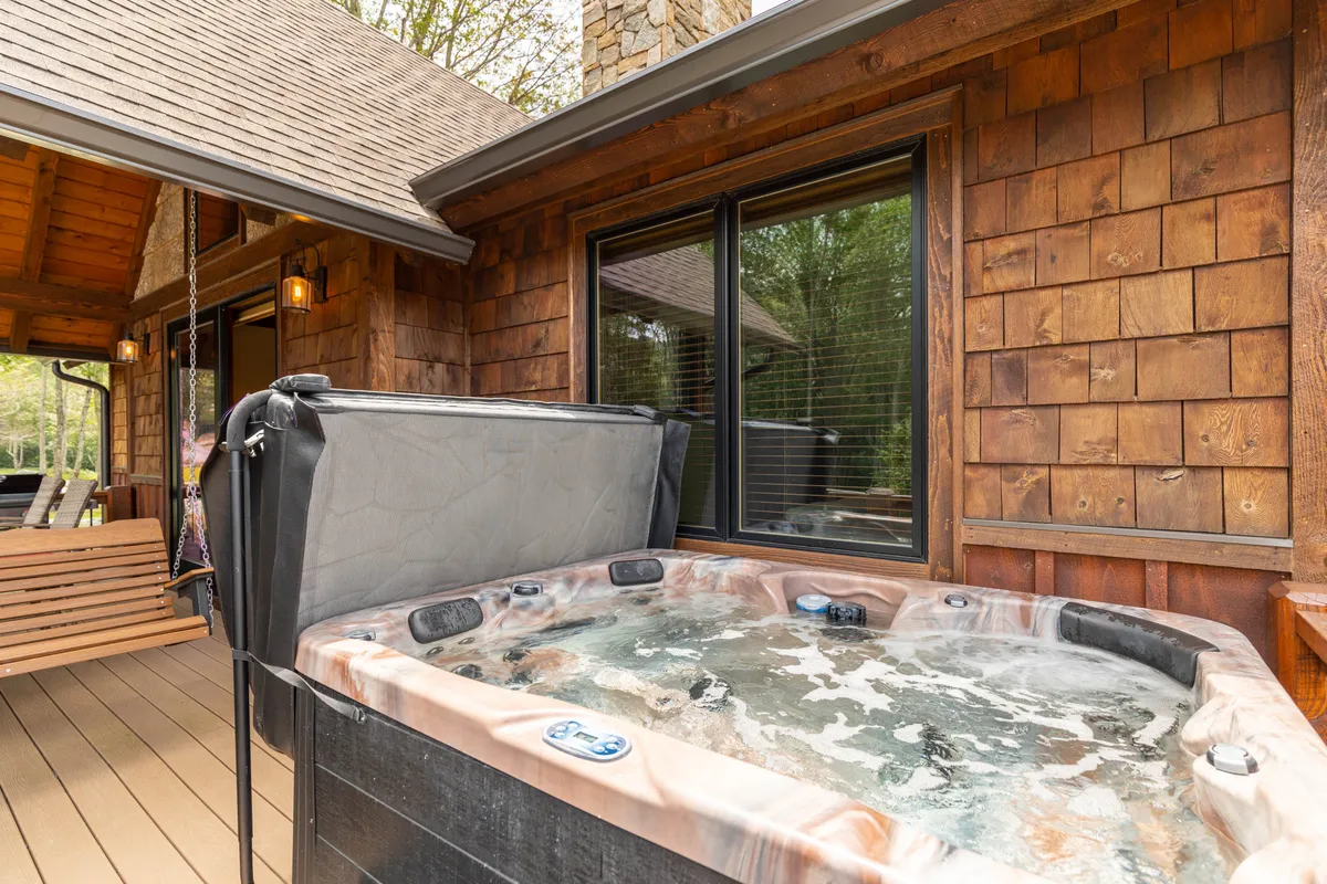 A tranquil exterior scene featuring a wooden deck with a bubbling hot tub, stone chimney, and large windows looking into the home. The hot tub sits prominently on the deck, surrounded by natural beauty, suggesting a peaceful spot for relaxation while enjoying the mountain air.