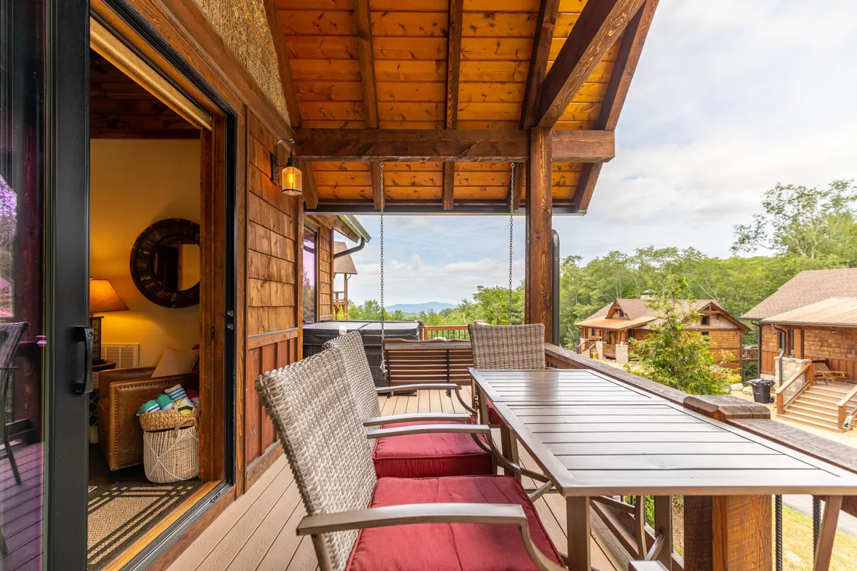 A cozy wooden balcony with a table and red cushions overlooks trees and mountains. The view through the door hints at the inviting interior, while the setup invites outdoor dining, relaxing, and socializing with nature as a backdrop.
